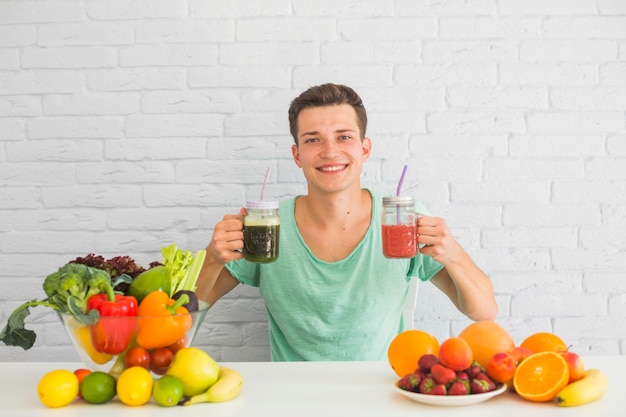 Foto gratuita hombre joven que sostiene el smoothie verde y rojo en su mano con la comida sana en la tabla