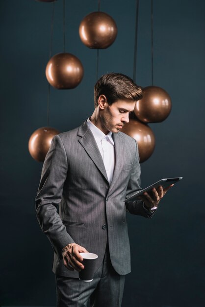 Hombre joven que sostiene la taza de café disponible que mira la tableta digital que se coloca delante del espejo aerodinámico alrededor de la lámpara de cobre