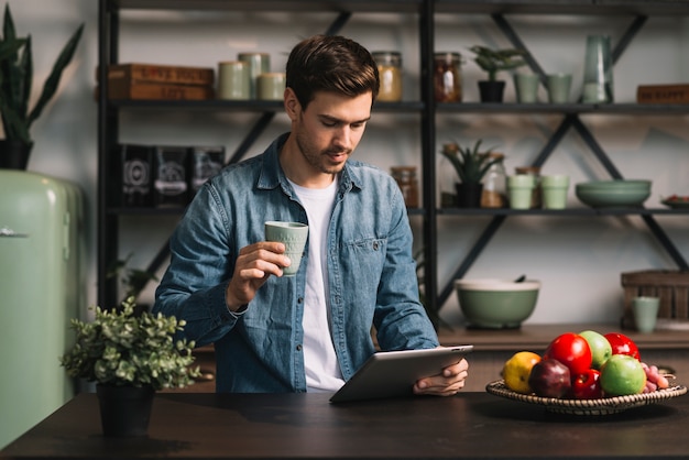 Foto gratuita hombre joven que sostiene la taza de café que mira la tableta digital