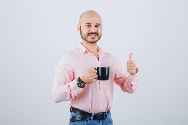 Hombre joven que sostiene la taza mientras muestra el pulgar hacia arriba en camisa rosa, vista frontal de los pantalones vaqueros.