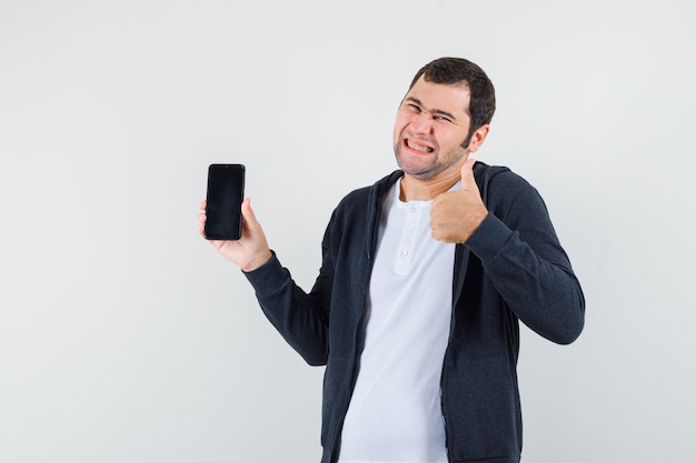 Hombre joven que sostiene el teléfono inteligente, mostrando el pulgar hacia arriba con una camiseta blanca y una sudadera con capucha negra con cremallera frontal y luciendo feliz. vista frontal.