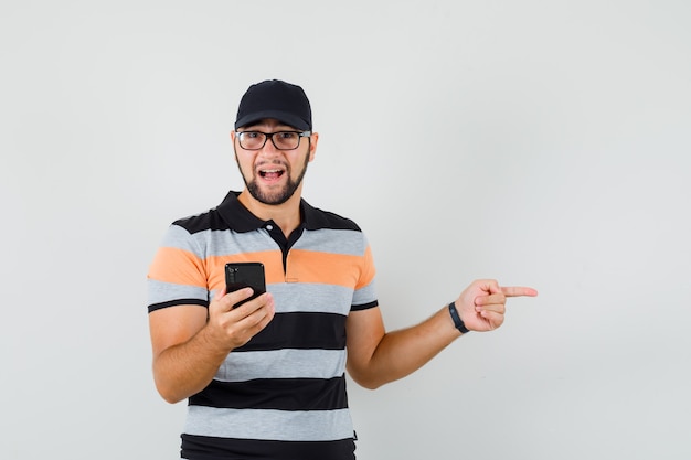 Hombre joven que sostiene el teléfono móvil, apuntando hacia el lado en camiseta, gorra y mirando positivo, vista frontal.