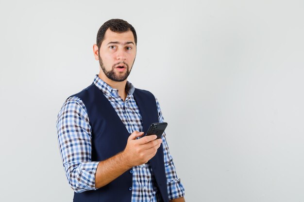 Hombre joven que sostiene el teléfono móvil en camisa, chaleco y mirando pensativo.