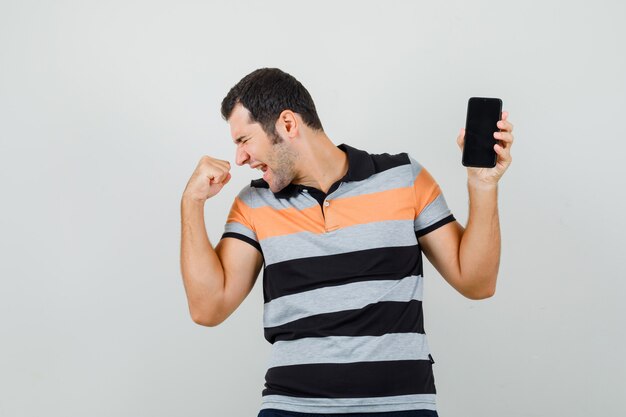 Hombre joven que sostiene el teléfono móvil mientras muestra el gesto del ganador en la camiseta y parece enérgico.