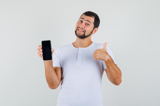 Hombre joven que sostiene el teléfono móvil mientras muestra el pulgar hacia arriba en la camiseta blanca y parece feliz. vista frontal.