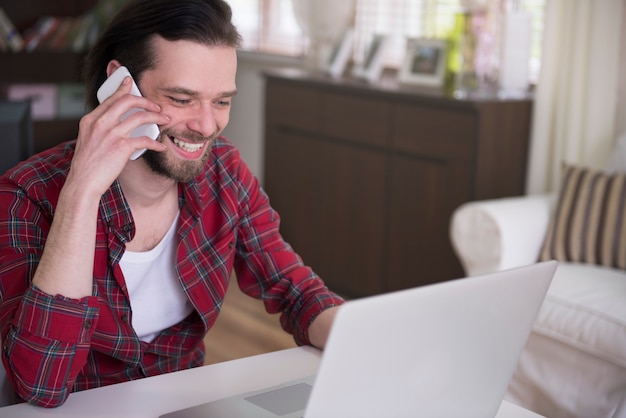 Foto gratuita hombre joven que trabaja en casa con su computadora portátil y hablando por teléfono