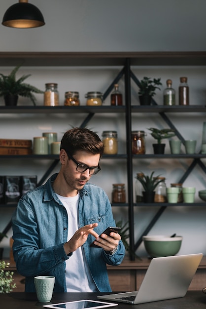 Foto gratuita hombre joven que usa el teléfono celular con la computadora portátil; tableta digital y taza de café en el mostrador de la cocina