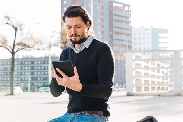 Hombre joven que usa el teléfono elegante que se sienta en banco contra el edificio de la ciudad