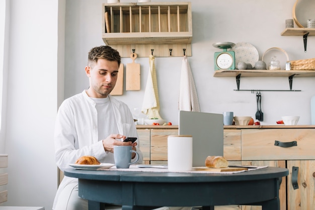 Hombre joven que usa el teléfono inteligente sentado cerca de la mesa con desayuno y una computadora portátil en la cocina