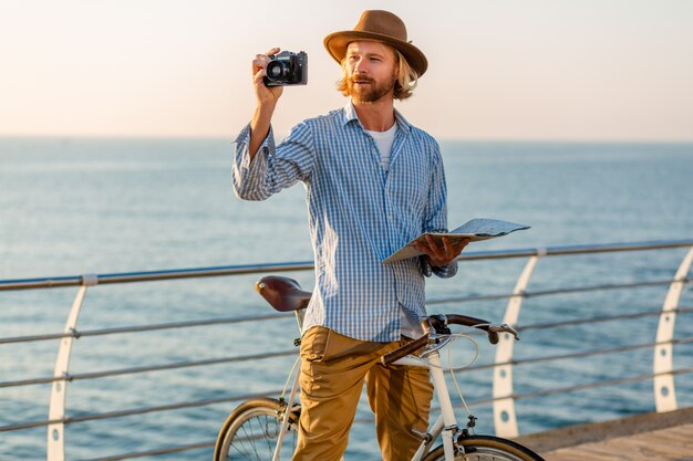 Hombre joven que viaja en bicicleta por el mar en las vacaciones de verano junto al mar al atardecer, sosteniendo el mapa de turismo tomando fotos en la cámara