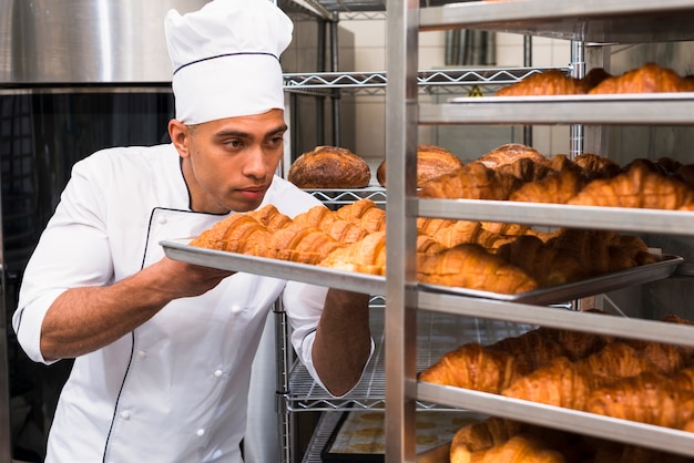 Hombre joven quitando la bandeja de croissant para hornear del estante