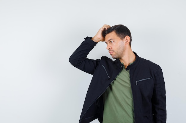 Hombre joven rascándose la cabeza mientras mira a un lado en camiseta, chaqueta y se ve guapo, vista frontal.