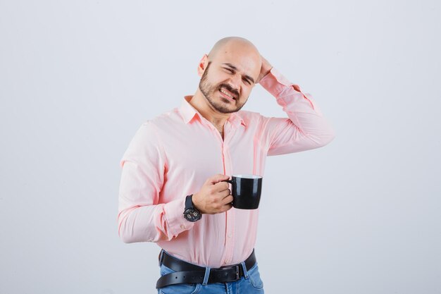 Hombre joven rascándose la cabeza mientras sostiene la taza en camisa rosa, jeans y mirando pensativo, vista frontal.