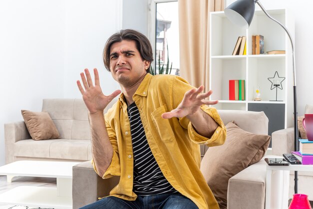 Hombre joven en ropa casual con expresión de disgusto haciendo gesto de defensa con las manos sentado en la silla en la sala de estar luminosa