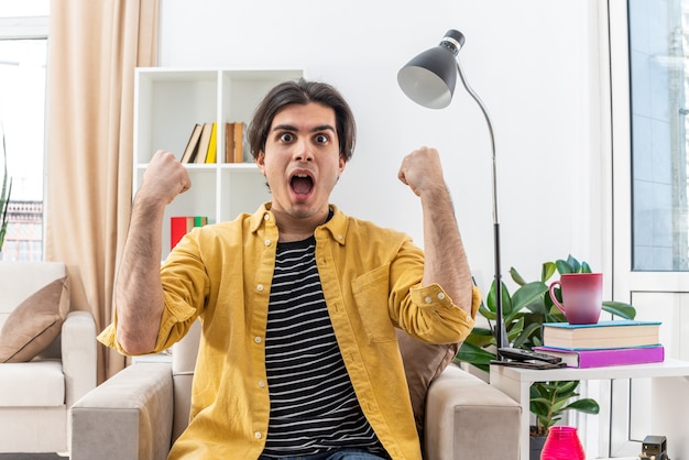 Hombre joven en ropa casual feliz y emocionado apretando los puños sentado en la silla en la sala de luz