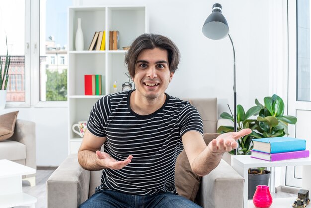 Hombre joven en ropa casual feliz y emocionado con los brazos extendidos sentado en la silla en la sala de luz