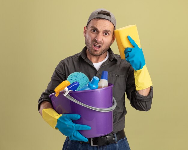 Hombre joven con ropa casual y gorra en guantes de goma sosteniendo un balde con herramientas de limpieza y una esponja que parece confundido y disgustado de pie sobre la pared verde