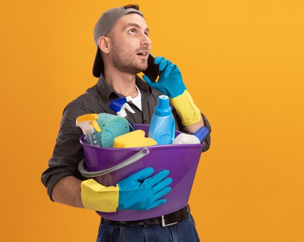 Hombre joven con ropa casual y gorra en guantes de goma sosteniendo un balde con herramientas de limpieza mirando hacia arriba sonriendo confiado mientras habla por teléfono móvil de pie sobre la pared naranja