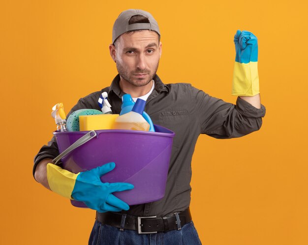 Hombre joven con ropa casual y gorra en guantes de goma sosteniendo un balde con herramientas de limpieza mirando con cara seria levantando el puño de pie sobre la pared naranja