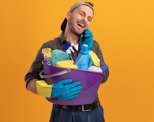 Hombre joven con ropa casual y gorra en guantes de goma sosteniendo un cubo con herramientas de limpieza sonriendo feliz y alegre mientras habla por teléfono móvil de pie sobre la pared naranja