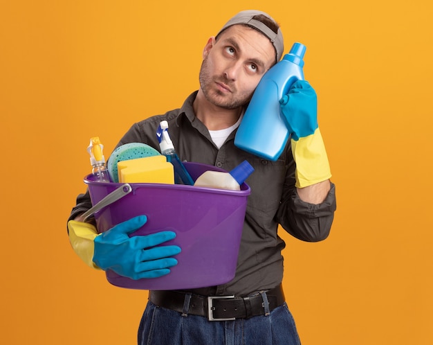 Hombre joven con ropa casual y gorra en guantes de goma sosteniendo un cubo con herramientas de limpieza y suministros de limpieza de botellas mirando hacia arriba desconcertado parado sobre la pared naranja