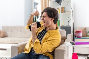 Foto gratuita hombre joven en ropa casual gritando estar enojado mientras habla por teléfono móvil apretando el puño sentado en la silla en la sala de luz