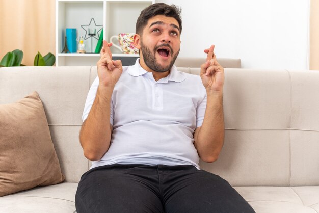 Hombre joven en ropa casual haciendo deseo deseable cruzar los dedos con expresión de esperanza sentado en un sofá en la sala de luz