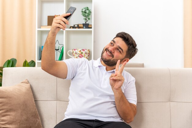 Hombre joven en ropa casual haciendo selfie con smartphone feliz y positivo mostrando v-sign sonriendo sentado en un sofá en la sala de luz