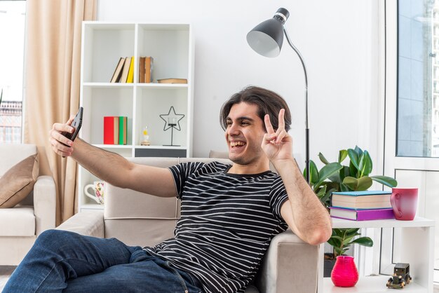 Hombre joven en ropa casual haciendo selfie con smartphone mostrando signo v feliz y alegre sentado en la silla en la sala de luz