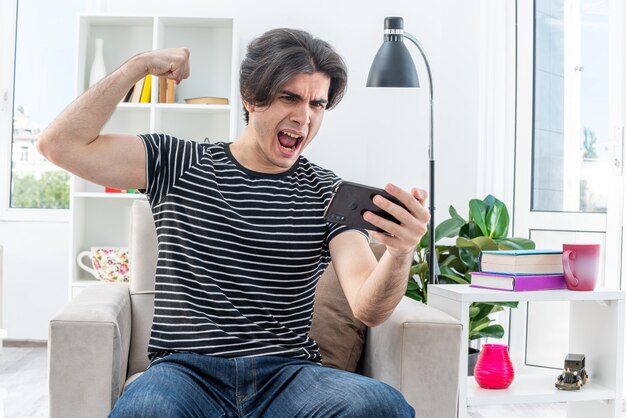 Hombre joven en ropa casual jugando con smartphone feliz y emocionado levantando el puño como un ganador sentado en la silla en la sala de luz