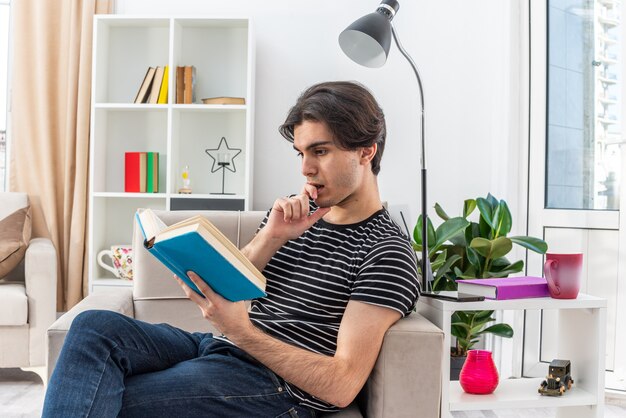 Hombre joven en ropa casual leyendo un libro con rostro serio sentado en la silla en la sala de luz