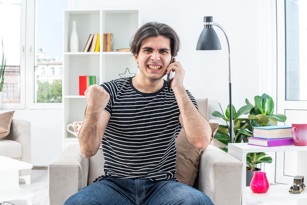 Hombre joven en ropa casual mirando con cara enojada mostrando el puño mientras habla por teléfono móvil volviéndose loco sentado en la silla en la sala de estar ligera