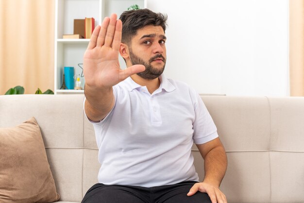 Hombre joven en ropa casual mirando con cara seria haciendo gesto de parada con la mano sentada en un sofá en la sala de luz