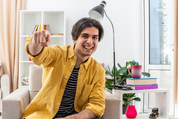 Hombre joven en ropa casual mirando feliz y alegre apuntando con el dedo índice al frente, sonriendo ampliamente sentado en la silla en la sala de luz