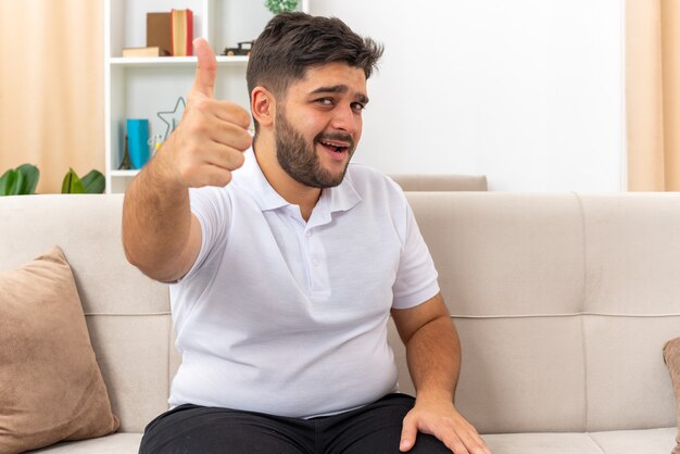 Hombre joven en ropa casual mirando feliz y alegre mostrando los pulgares para arriba sentado en un sofá en la sala de luz