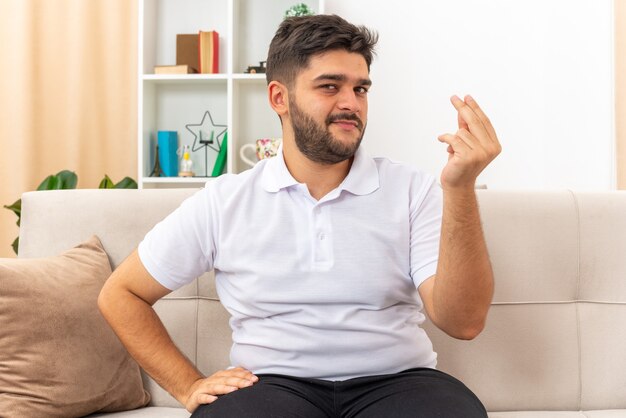 Hombre joven en ropa casual mirando hacer gesto de dinero frotando los dedos sonriendo astutamente sentado en un sofá en la sala de luz