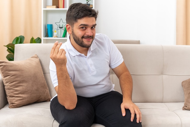 Hombre joven en ropa casual mirando hacer gesto de dinero frotando los dedos sonriendo astutamente sentado en un sofá en la sala de luz