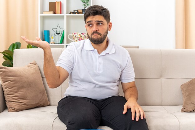 Hombre joven en ropa casual mirando a un lado confundido presentando algo con el brazo de la mano sentado en un sofá en la sala de estar luminosa