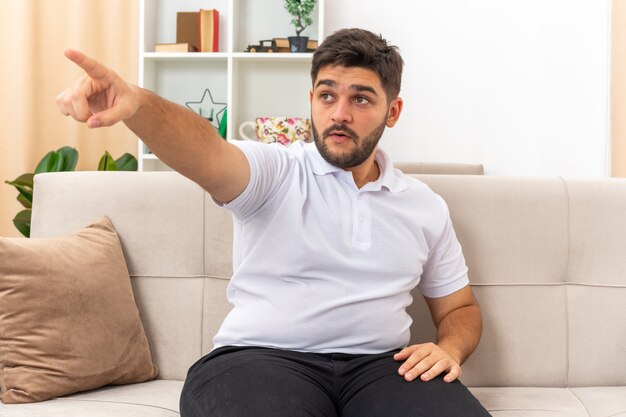 Hombre joven en ropa casual mirando a un lado sorprendido apuntando con el dedo índice hacia el lado sentado en un sofá en la sala de luz