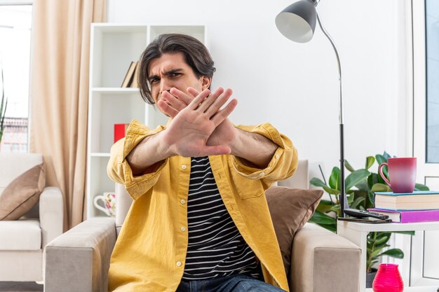 Hombre joven en ropa casual preocupado haciendo gesto de parada con las manos cruzando los brazos sentado en la silla en la sala de luz