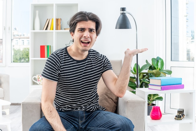 Hombre joven en ropa casual que parece confundido y disgustado levantando el brazo con disgusto e indignación sentado en la silla en la sala de estar luminosa