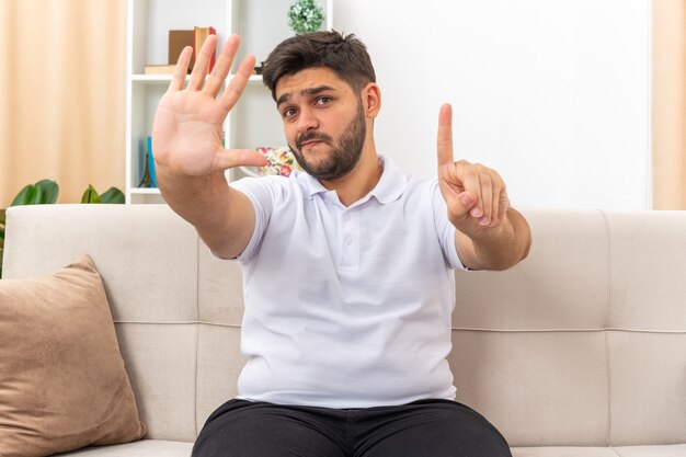 Hombre joven en ropa casual que parece confundido y disgustado mostrando el número seis sentado en un sofá en la sala de estar luminosa