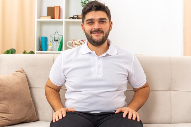 Hombre joven en ropa casual que parece feliz y positivo pasar el fin de semana en casa sentado en un sofá en la sala de luz