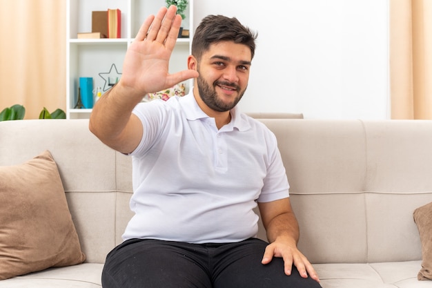 Hombre joven en ropa casual que parece feliz y positivo saludando con la mano feliz y positivo sentado en un sofá en la sala de luz