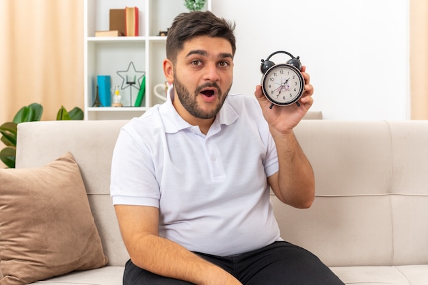 Hombre joven en ropa casual con reloj despertador feliz y sorprendido sentado en un sofá en la sala de luz