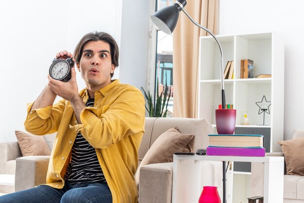 Hombre joven en ropa casual con reloj despertador mirando asombrado y sorprendido sentado en la silla en la sala de luz