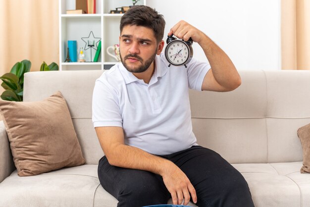 Hombre joven en ropa casual con reloj despertador mirando a un lado con expresión escéptica sentado en un sofá en la sala de estar ligera