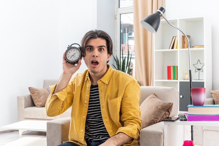 Foto gratuito hombre joven en ropa casual con reloj despertador mirando sorprendido sentado en la silla en la sala de luz