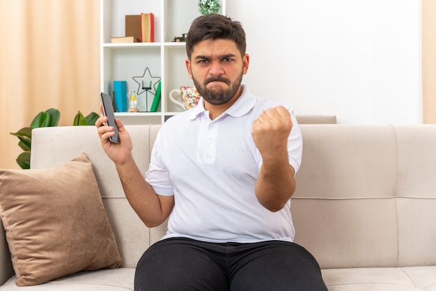 Hombre joven en ropa casual con smartphone apretando el puño mirando enojado y frustrado sentado en un sofá en la sala de luz