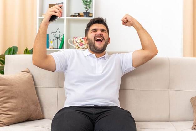 Hombre joven en ropa casual con smartphone feliz y emocionado apretando el puño regocijándose de su éxito sentado en un sofá en la sala de estar luminosa